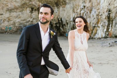 A happy wedding couple runs on the beach at Shark Fin Cove in California.