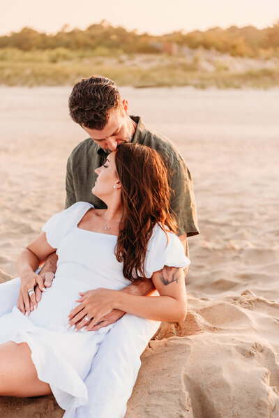 a man and woman sitting on the sand at the beach during their engagement session.  The man is kissing his fiances head, while she has her hands over his.  photo taken by Cape May photographer, Kristi