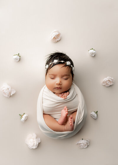 cute baby sleeping on white blanket with flowers around her wearing a flower headband