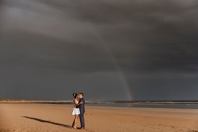 Two people getting married and hugging at The Dunes with a cloudy sky and rainbow in the background