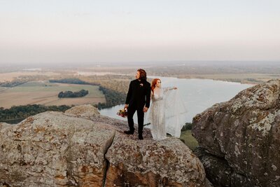 An eloping couple stands on a large boulder atop the cliffside of Petit Jean State Park in Arkansas. The bride is in a  silver dress that is adorned in sequins that make it look like the night sky full of twinkling stars. She holds hands with her groom who is in a full black suit and is holding her colorful bouquet in his other hand. A view of the Arkansas River and valley can be seen in the background.