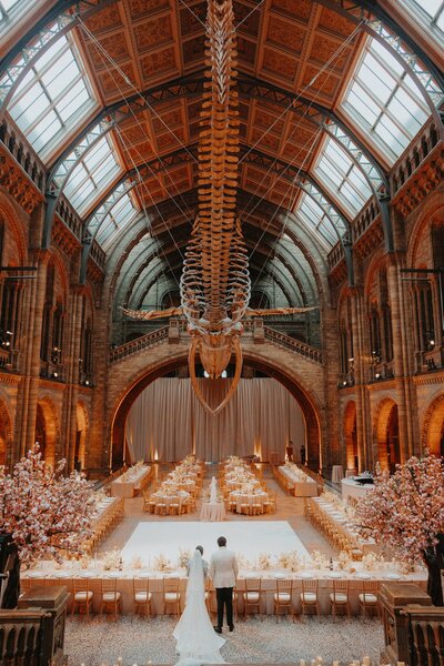 Bride and groom standing hand-in-hand in the Hintze Hall of the Natural History Museum, London, overlooking a luxury wedding setup featuring round tables.