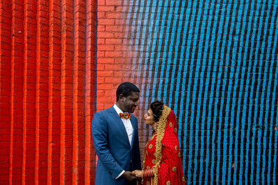 A couple holding hands and looking at each other while standing in front of a colored wall.