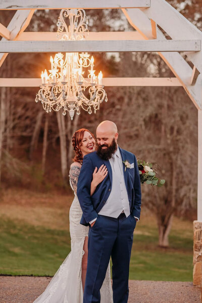 Photo of a bride cuddling a grooms back and laughing underneath a chandelier outside