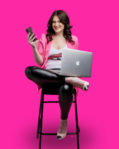 Emily sitting on a tall black chair, holding her laptop on her leg and holding a phone in the other hand. She is wearing a pink blazer and smiling at the camera in front of a hot pink background.