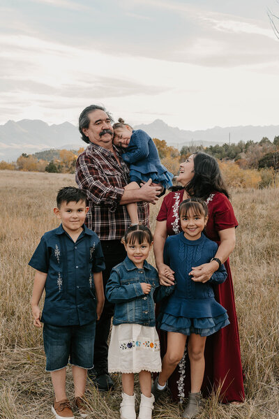 Playful family photos in Montrose Colorado show a family of five posing in a field with mountains in the background, all smiling and dressed in blue and red.