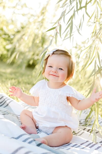 Infant sits on a blanket among willow branches for family photography shoot.
