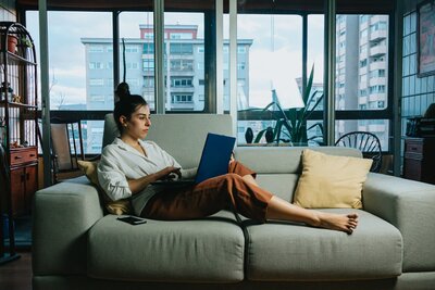 Woman in orange pants and white shirt laying on a couch in front of windows looking at her laptop