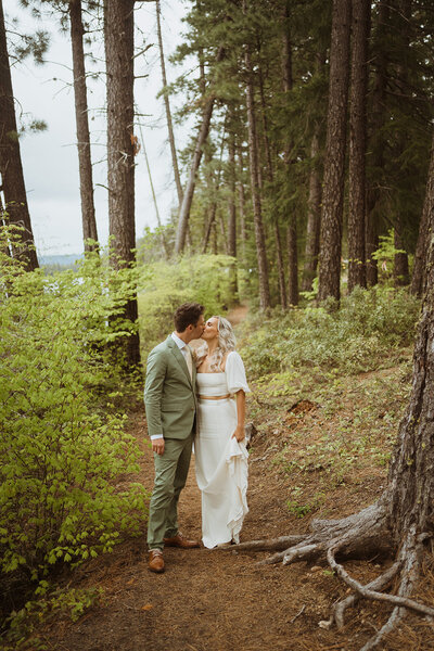 bride and groom are stopped in the middle of the hiking trail kissing. the bride is lifting her dress slightly, and the groom is in a forest green suit. they are in the middle of the woods surrounded by tall trees and greenery.