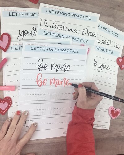 Hand lettering practice sheets with "be mine" in red and black and hands writing