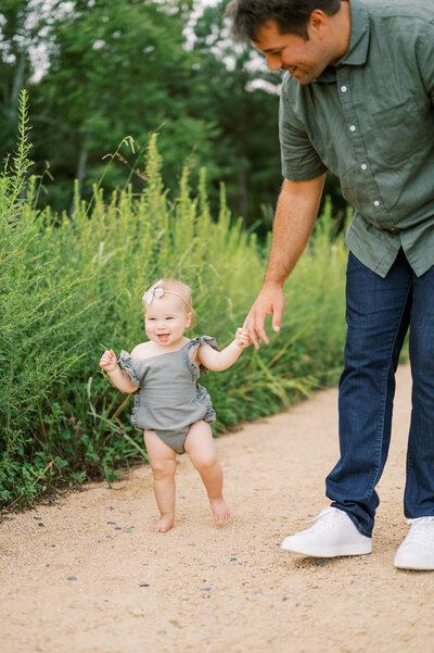 Dad holds hand of one year old baby girl in green romper during outdoor family photos