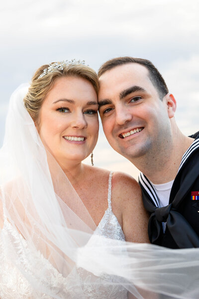 Bride and Groom kissing on a glass walkway in urban Norfolk Waterside district.  Beautiful lines and framing.  Small amount of flash light on front of Bride and Groom.  Bride holding bouquet.  Dressed in white  strappy dress with mid length veil, holding a bouquet of florals.  Groom in navy dress uniform.