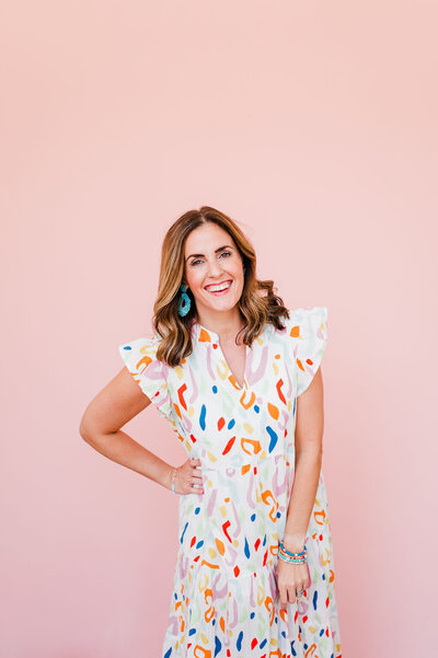 female photographer in multi color dress standing against a pink backdrop