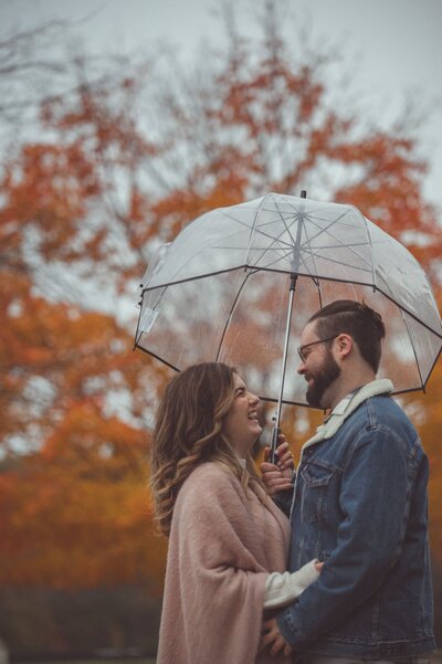 Engagement Couple Photo, the couple are standing under an umbrella, in a gloomy Fall afternoon, with a Fall colored trees in the backdrop