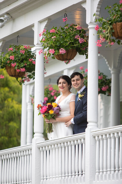 a bride and groom stand on a porch with white pillars decorate with pink flowers Mountainville Manor wedding photographer Milestone Images