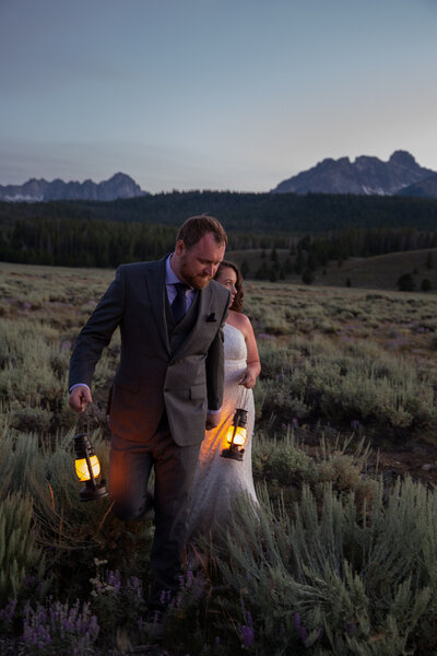 A bride and groom walk through a meadow holding lanterns during the sunset in Idaho.