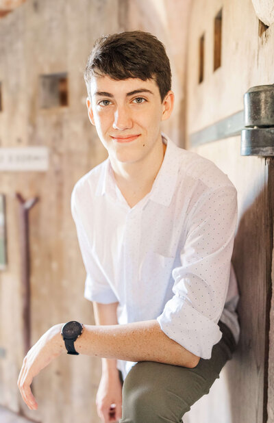 A young person with short dark hair, wearing a white button-up shirt and a black watch, poses indoors for a senior photographer in Fayetteville, NC. They lean slightly forward with a neutral expression against the backdrop of a wooden surface with a latch.