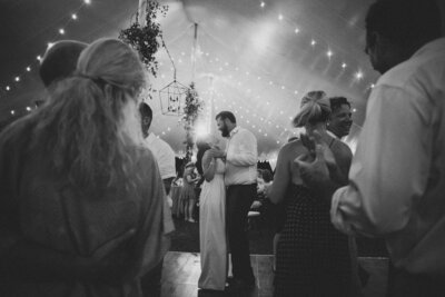 a bride and groom dance under a tented wedding reception and are surrounded by friends and string lights