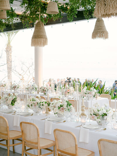 Outdoor wedding reception set up under an awning with straw chandeliers hanging from it. The white table clothed table is decorated with light pink and white flowers