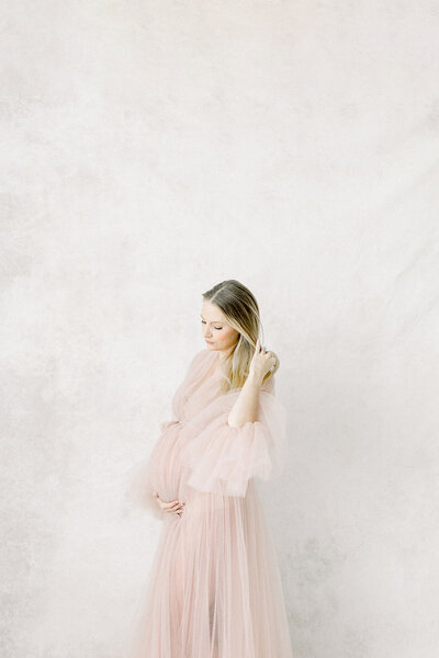 A photo of a pregnant woman dressed in a long blush tulle gown holding her belly while standing by the wall in a Dallas/Fort Worth photography studio.