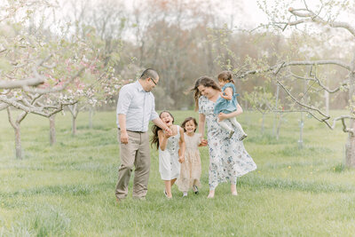 A mother and father and their three daughters hold hands and are walking through an apple orchard while giggling