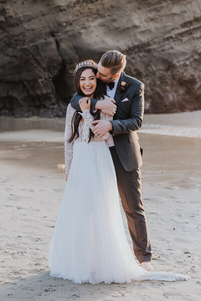 Wedding Photographer, groom kisses bride on side of head, they stand at the beach