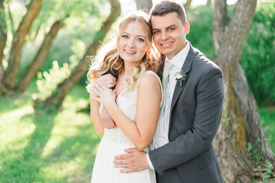 Groom with his arms around the bride in a willow grove with the sun shining on them.