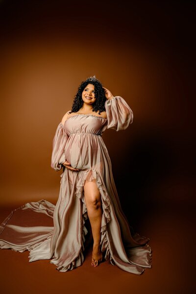 Charlotte Area Mom dressed in a golden brown dress on a Studio background with one hand cradling her bump her leg exposed and the other hand touching a crown on her head