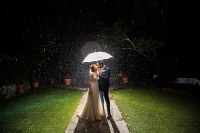 Bride and Groom stand in the rain under an umbrella sharing a kiss