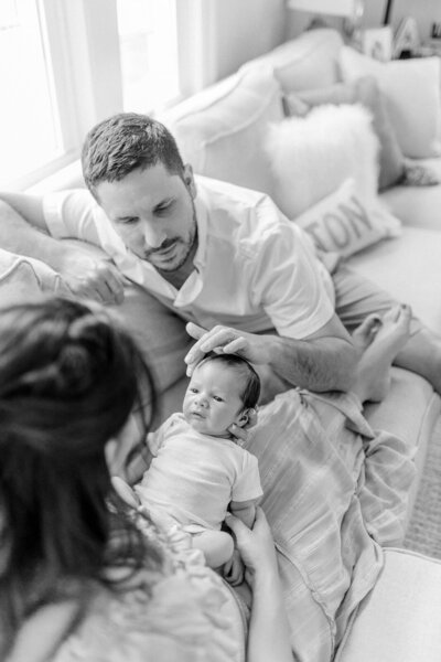 A black and white image of a mother holding her newborn baby boy on her lap while the baby's father brushes his hair with his finger