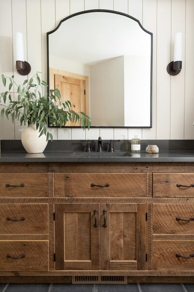 Large rustic wood vanity with lots of drawers, and dark grey countertops. Wall behind the vanity is off-white shiplap with a large arched mirror and two oil rubbed bronze sconces on either side. A widespread oil rubbed bronze faucet at the sink with a large beige vase with green florals to the left of the sink. To the right of the sink a jar of hand soap and a small beige dish. Large rustic black square floor tiles with brown grout.