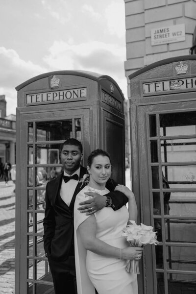 groom leans against a london telephone box with his arm around his bride who leans back on him whilst holding a bouquet of calla lilies