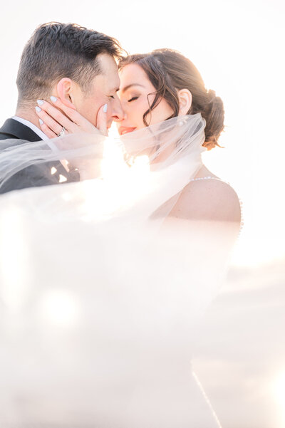 A loving couple kissing as her wedding veil sweeps across her face at sunset by destination wedding photographer, JoLynn Photography