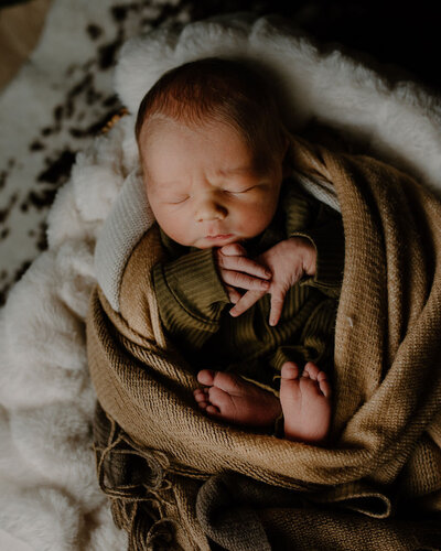 Adorable vintage looking newborn photoshoot by Morgan Ashley Lynn Photography in Milwaukee, WI of a baby boy in an olive green matching set wrapped up in a brown blanket with his hands touching and feet up, sleeping. He is laying on a soft bulky white blanket and on a rug.
