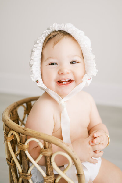 Baby girl in white bonnet smiles  during baby photography session in Raleigh NC studio