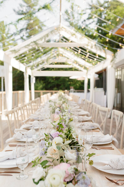 Wedding reception set up in a clear canopy with twinkly lights, white dishes, bronze cutlery, and light purple, pink, and white flowers