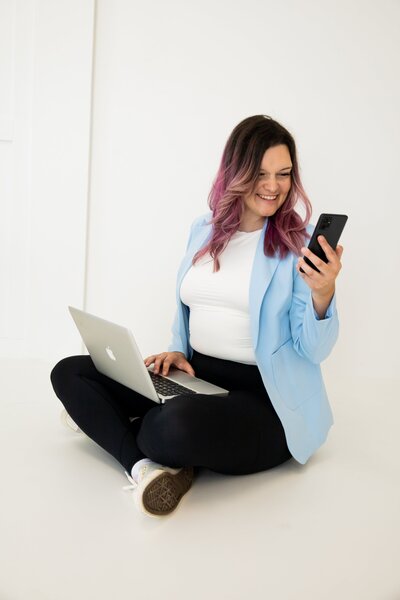 Founder of Florida Social Butterfly in blue blazer sitting cross legged with laptop and phone checking on social media clients