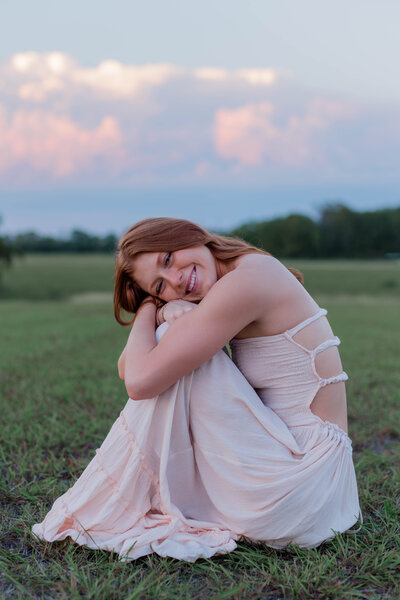 Red-headed senior girl poses in a shy pose while hugging her knees in a green meadow at sunset