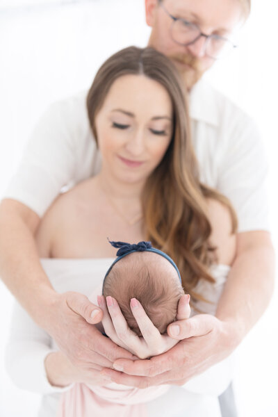 A mother and father smile down at their newborn baby in their hands in a bow