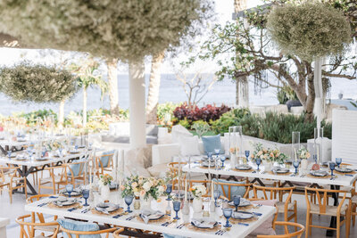 Outdoor wedding reception set up with a white table decorated with blue glasses, gold charger plates, and white and peach flowers.