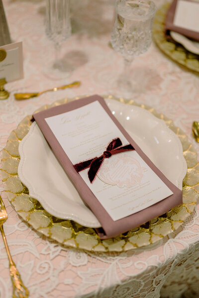 Exquisite place setting featuring a gold charger, blush napkins, and a gold foil menu paired with a blush die-cut card and burgundy velvet ribbon.