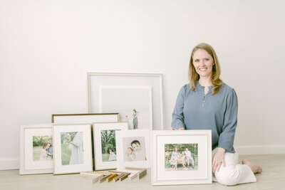A picture of Molli Dill sitting on the floor holding a framed family portrait with other family portraits next to her and frame corners beside her