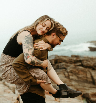 Girl riding piggy back on guy on rocky shore line with ocean waves in background