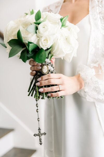 Close up detail of bride holding her white rose bouquet with a rosary tied around the stems