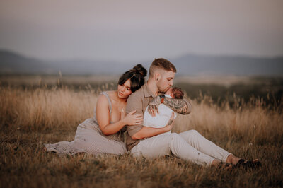 Couple sitting in a field . Dad is kissing newborn while mum rests against dad's back.
