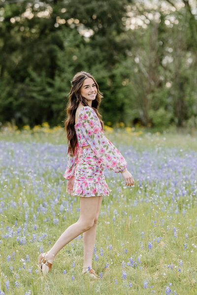 senior portrait of girl in floral print dress walking through a field of bluebonnets and wild flowers