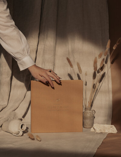 Hand reaching to  orange luxury wedding album  that is standing between two jars with cattail in them with a linen backdrop during the evening -Romero Album Design