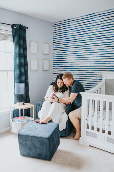 Couple smiles at newborn baby boy in blue and white nursery during in-home photography session