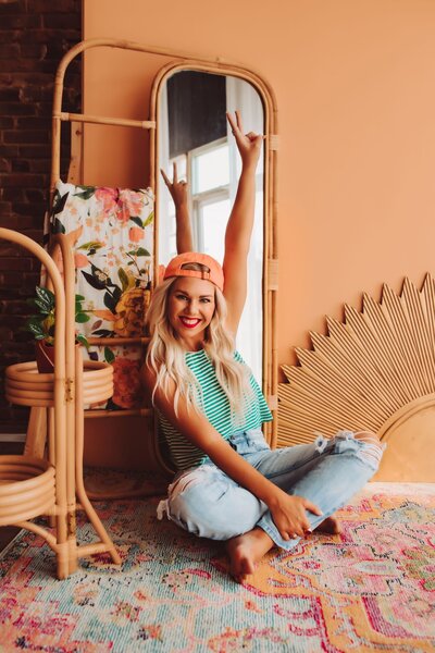 Founder of Infinite Productions, Andi Sweeny, sitting cross legged on a floor wearing ripped jeans, a blue and white striped shirt, and an orange hat raising her hand to give the peace sign