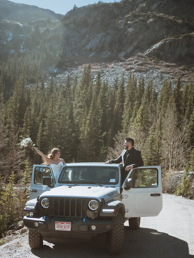 Couple standing out opposite doors of their jeep cheering and smiling at each other. the bride is holding her bouquet, and the groom is wearing a black suit. the jeep is silver. they are in the colorado mountains after their hiking elopement.
